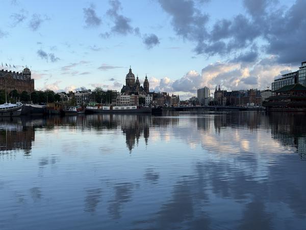 This photo captures a serene view of the Amsterdam skyline from Prins Hendrikkade, Centrum, with historic buildings and modern structures reflected in the calm water